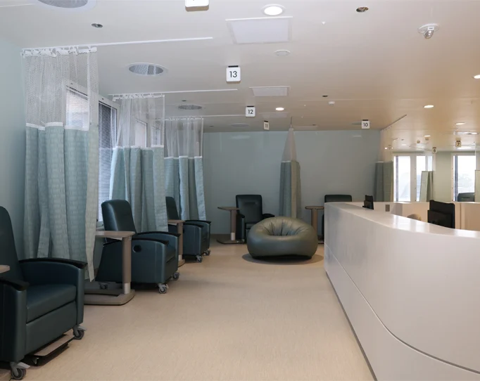 Modern hospital infusion room with reclining chairs, privacy curtains, a bean bag, small tables, and a curved white nurses’ station under bright overhead lights.