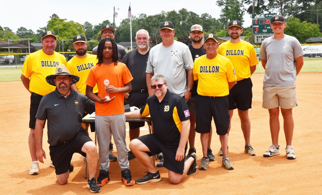 A group of men and a boy pose on a baseball field, with the boy holding a small trophy. Most adults wear yellow team shirts.