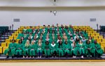 A large group of graduates wearing green caps and gowns pose for a class photo seated on bleachers in a gymnasium.