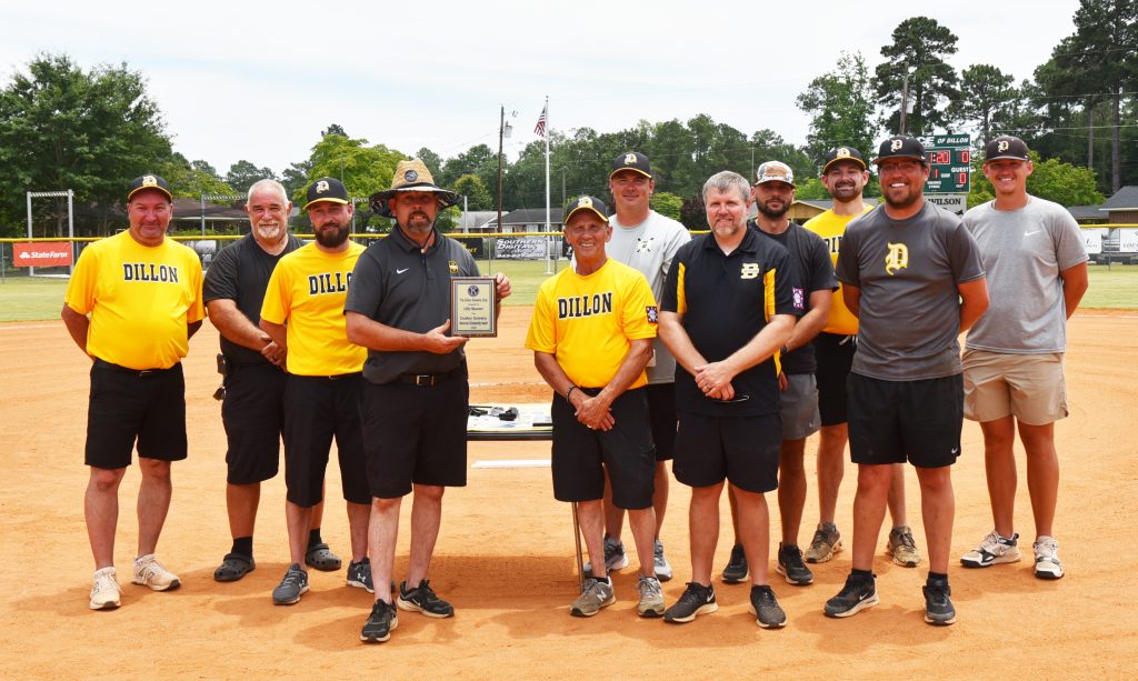 A group of men in sports attire stand on a baseball field, with one man holding a plaque. Trees and flags are visible in the background.