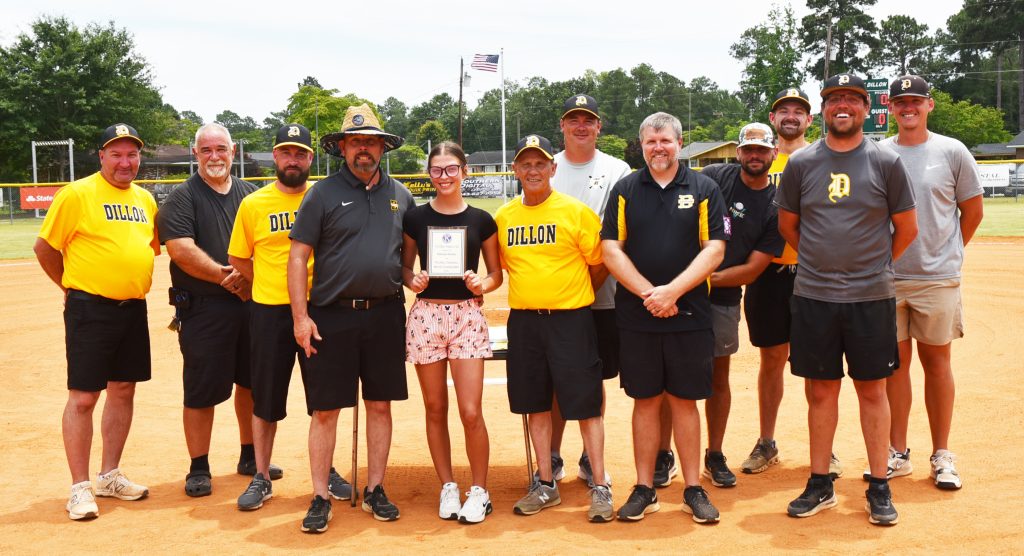 A group of people stands on a baseball field, with one person in the center holding a plaque and others smiling around them, wearing casual sports attire.