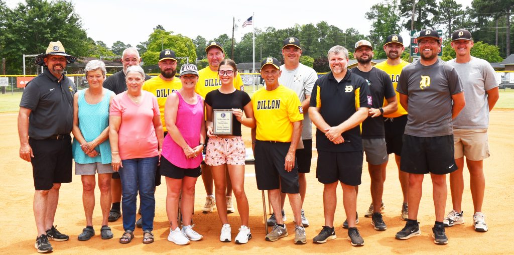 A group of people standing on a baseball field, one person in the center holding a plaque, some wearing yellow "BALCOY" shirts.