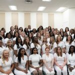 A large group of women dressed in white uniforms pose together in a well-lit indoor room, smiling and facing the camera.