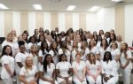 A large group of women dressed in white uniforms pose together in a well-lit indoor room, smiling and facing the camera.