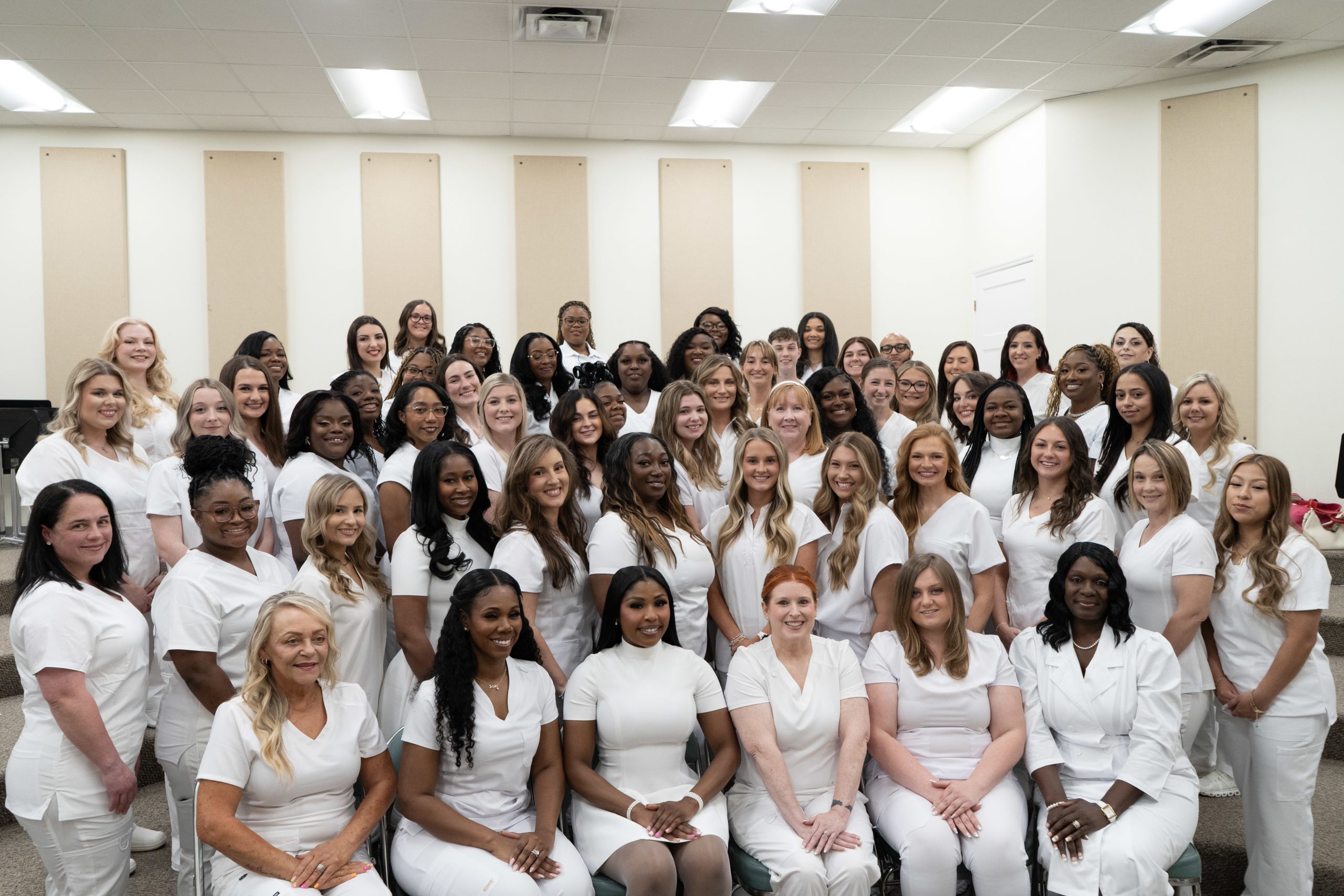 A large group of women dressed in white uniforms pose together in a well-lit indoor room, smiling and facing the camera.