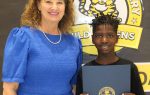 A woman in a blue dress stands next to a smiling boy holding a certificate in front of an elementary school sign with a wildcat logo.