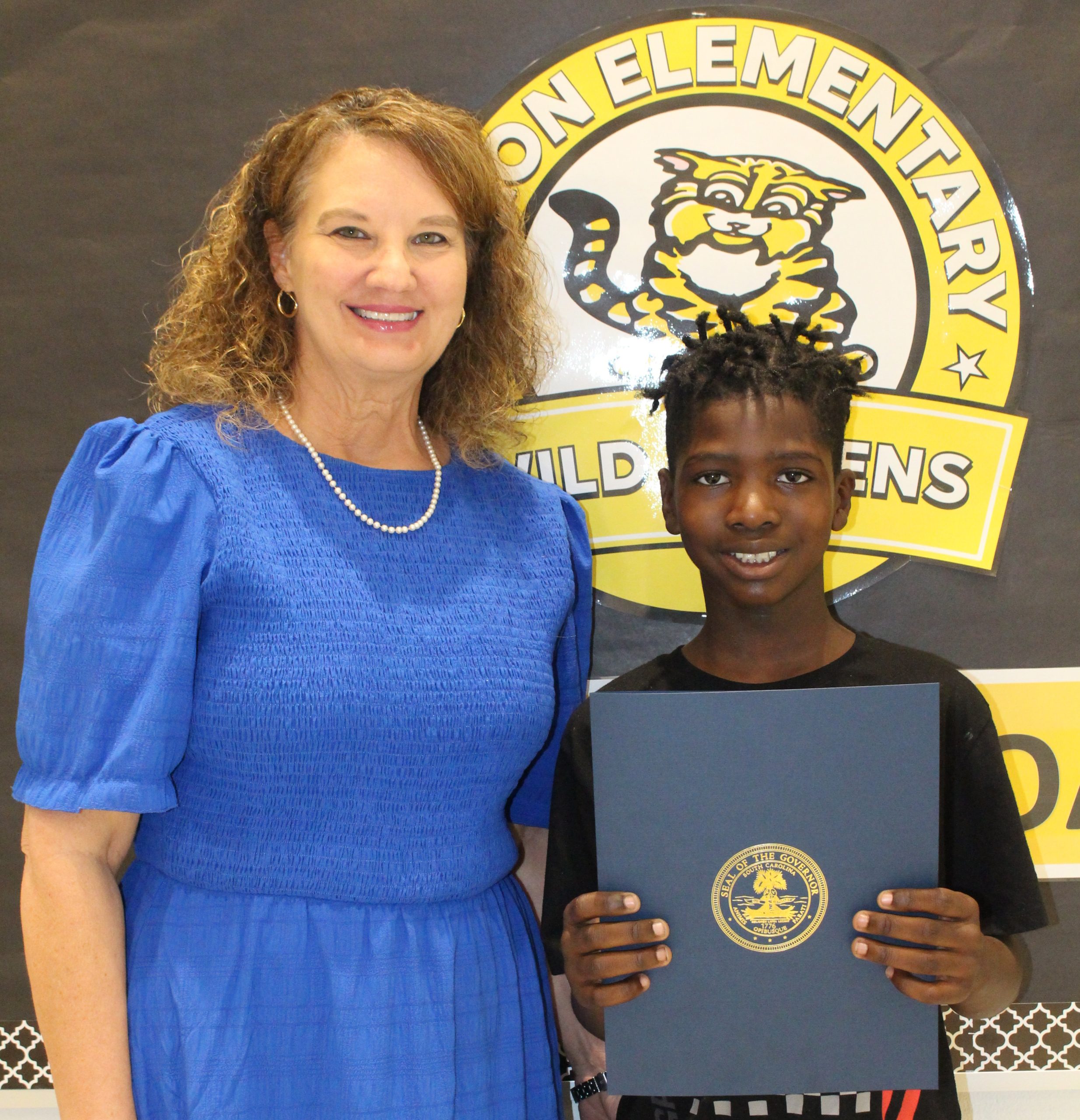 A woman in a blue dress stands next to a smiling boy holding a certificate in front of an elementary school sign with a wildcat logo.
