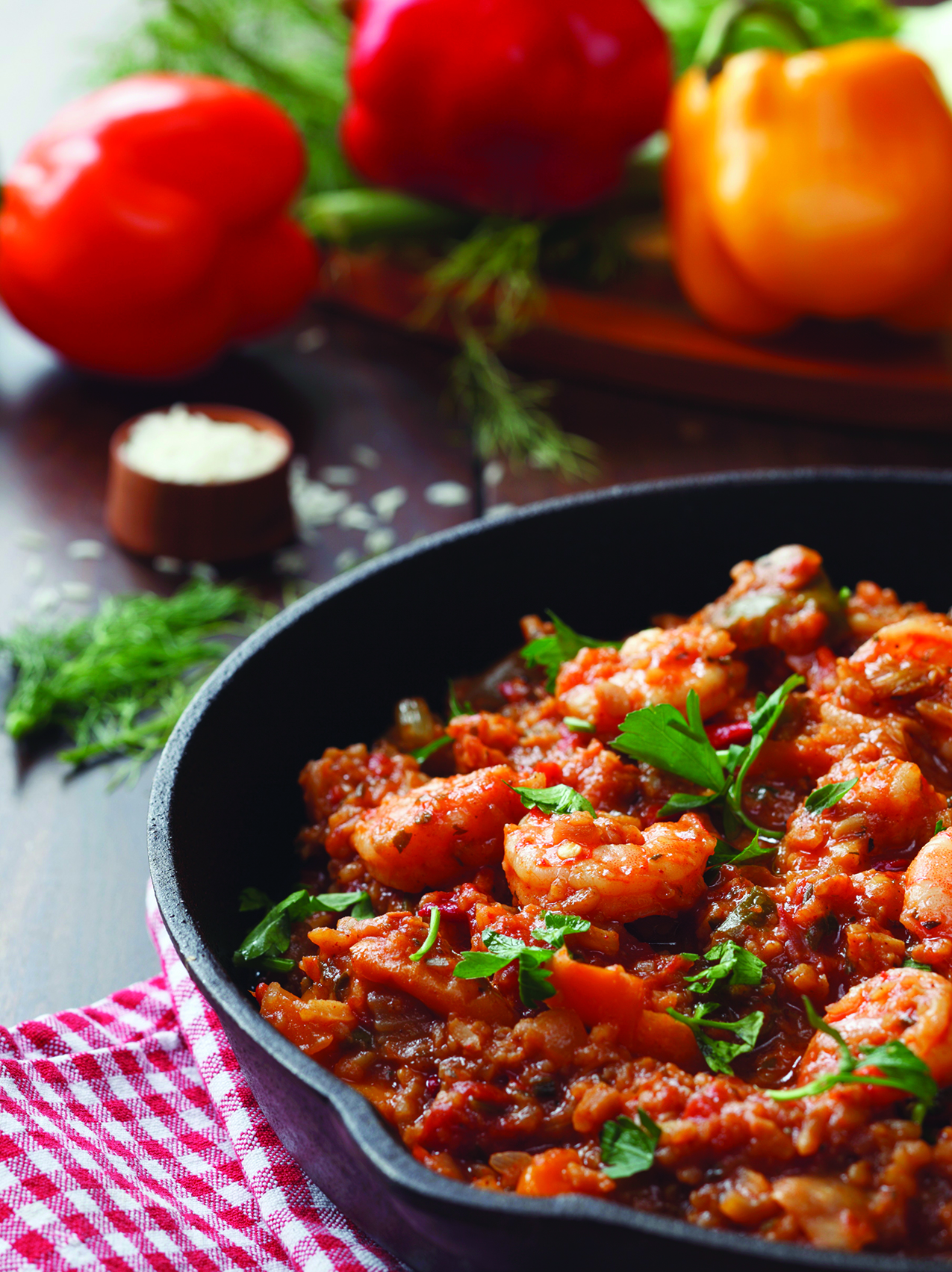 A close-up of shrimp jambalaya in a black skillet, garnished with fresh herbs, with bell peppers and rice in the background.