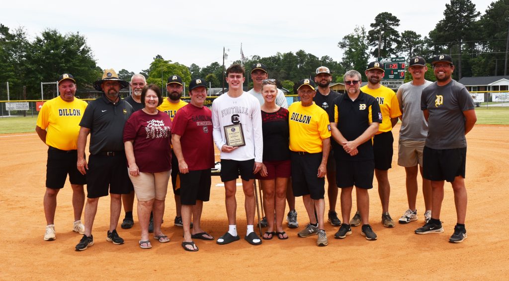 A group of people standing on a baseball field, one person in the center holding a plaque or award, smiling for the camera.
