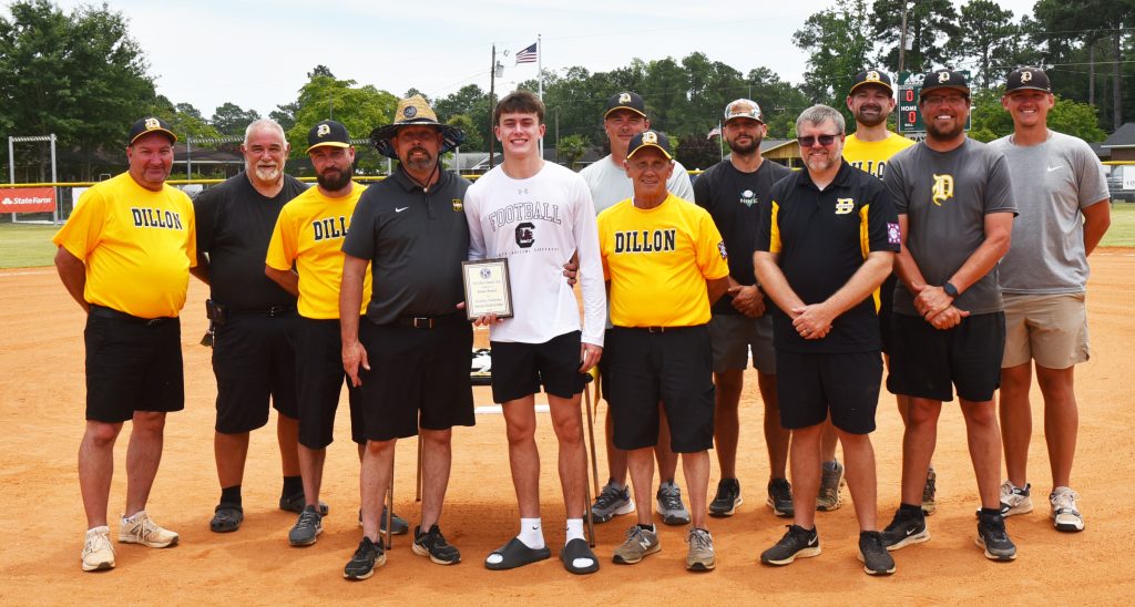 A group of men stand on a baseball field, with one young man in the center holding a plaque, surrounded by coaches in yellow and black shirts.