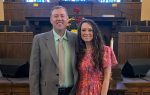 A man in a gray suit and a woman in a pink dress smile, standing together inside a church with wood paneling and pews.