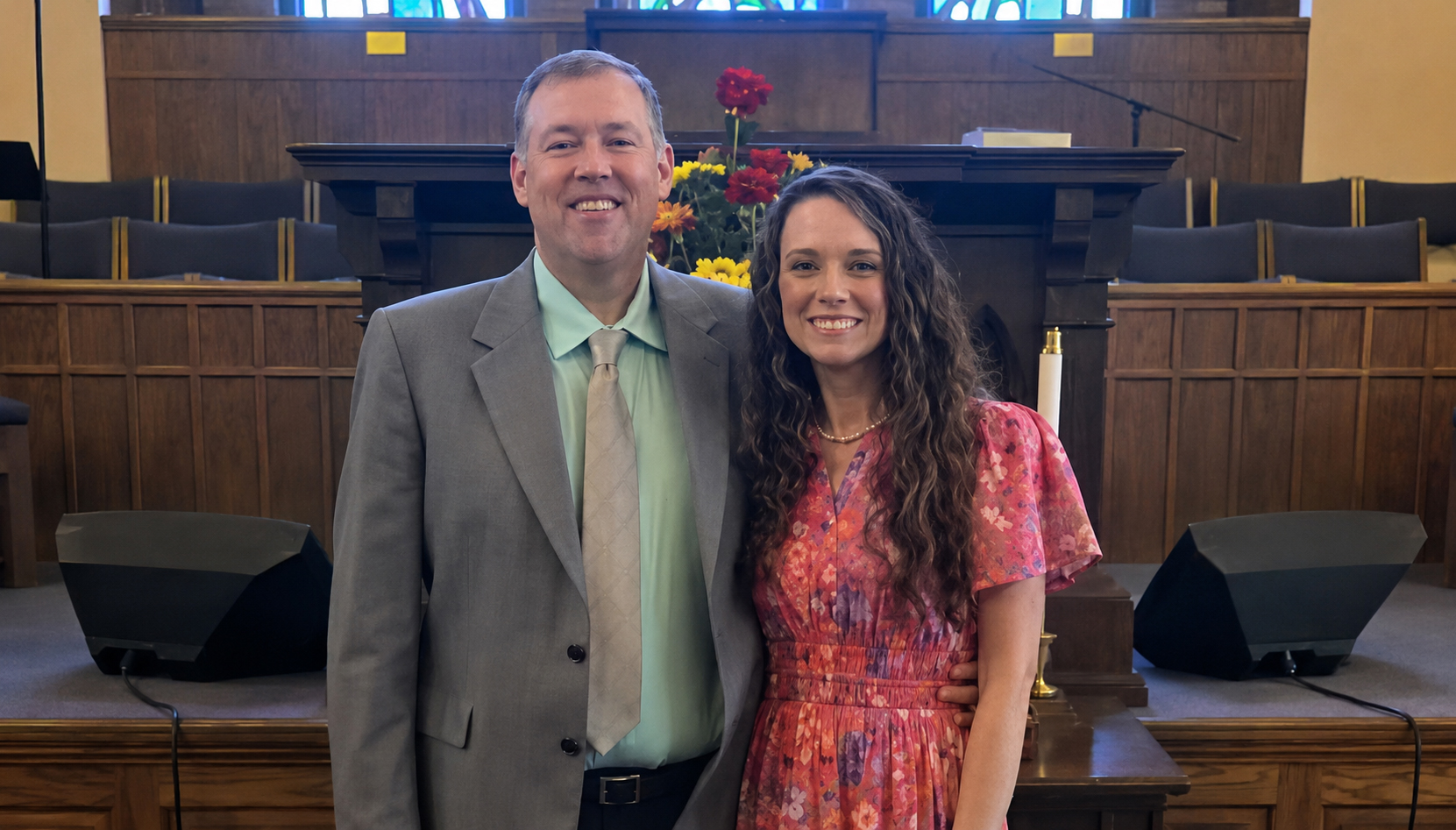 A man in a gray suit and a woman in a pink dress smile, standing together inside a church with wood paneling and pews.