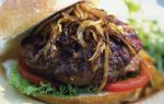 A hamburger topped with grilled onions, lettuce, and tomato, served in a sesame bun on a plate, with some salad in the background.