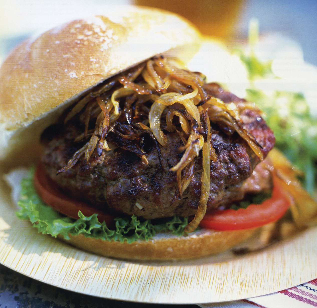 A hamburger topped with grilled onions, lettuce, and tomato, served in a sesame bun on a plate, with some salad in the background.
