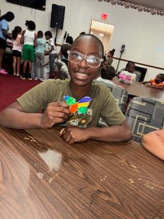 Smiling boy wearing glasses sits at a table holding a colorful butterfly craft, with other children and crafts visible in the background.