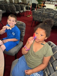 Two children sit on chairs indoors, smiling and holding small toys or snacks, with rows of patterned chairs and a red carpet visible in the background.