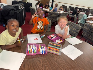 Three children sit at a table with markers and paper, smiling and posing for the camera while drawing in a classroom setting.