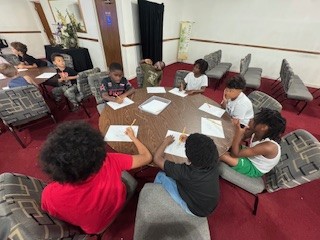 Children sitting around a round table, writing on paper, in a room with red carpeting and several chairs, suggesting a group activity or classroom setting.
