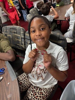 A young girl smiling and holding a craft project, sitting at a table with other children in a classroom or activity room.