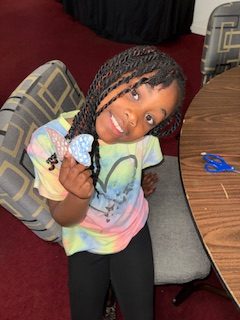 Smiling girl with braids in a tie-dye shirt holds butterfly crafts, sitting on a chair next to a table with blue scissors.