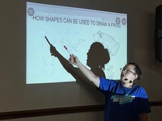 Person in blue shirt points at frog drawing tutorial projected on screen, explaining how shapes can be used to draw a frog.