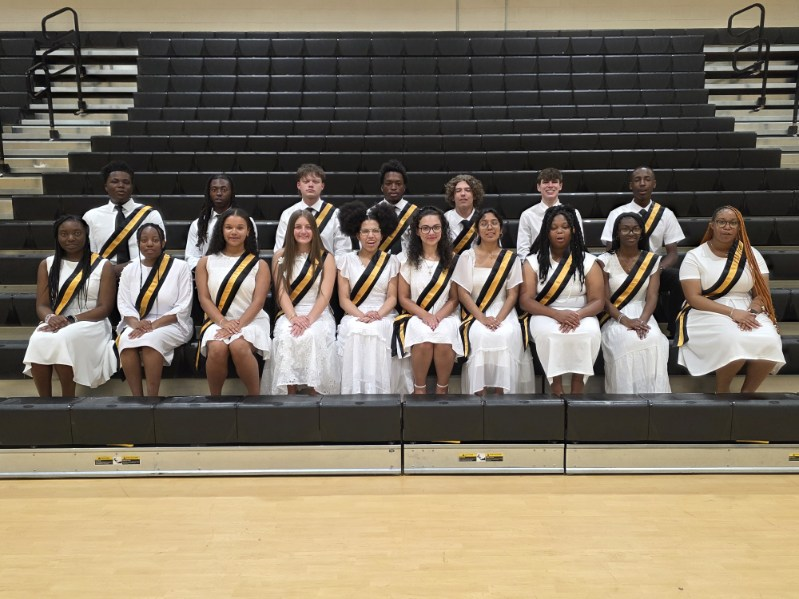 A group of students in white outfits with sashes sit and stand on bleachers in a gymnasium, posing for a formal group photo.