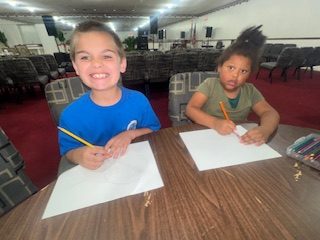 Two children sitting at a table, smiling and drawing on white paper with pencils in a large room filled with empty chairs.