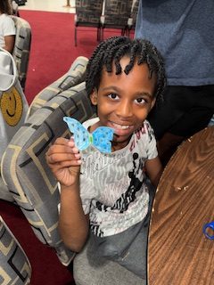 Smiling child sitting at a table, holding up a blue paper butterfly craft, with patterned chairs and a red carpet in the background.