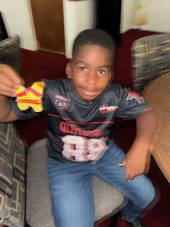 A young boy in a sports jersey sits on a chair, smiling and holding a decorated star-shaped cookie.