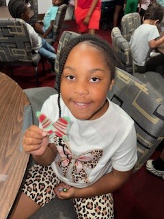 Smiling girl sitting at a table holds a small, striped craft project, wearing a white shirt with a bow and leopard print shorts, in a busy indoor setting.