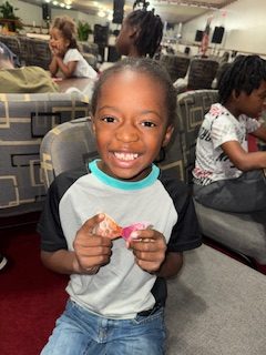 Smiling young boy sitting on a chair indoors, holding a small toy, with other children in the background.