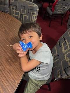 A young boy sitting at a table smiles while holding a blue toy or object, surrounded by patterned chairs in a room with a red carpet.