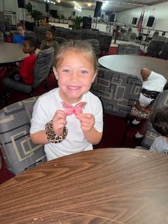 Smiling young girl sitting at a table indoors, holding a pink butterfly-shaped object, with other children and chairs in the background.
