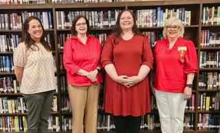 Four women smiling, standing in front of bookshelves in a library, three wearing red tops and one in a light patterned blouse.
