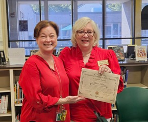Two women in red tops smiling and holding a certificate in a library setting, with bookshelves and large windows in the background.