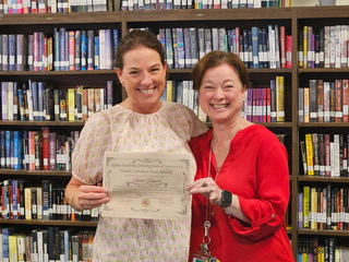 Two women smiling in a library, one holding an award certificate, standing in front of bookshelves filled with books.