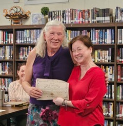 Two women smiling and holding a certificate together in a library, with bookshelves and another person visible in the background.