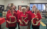 Seven women wearing red tops stand indoors, three in front holding large gold letters "D," "K," and "G" while smiling for the photo.