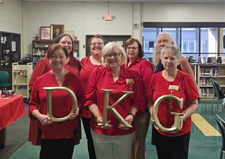 Seven women wearing red tops stand indoors, three in front holding large gold letters "D," "K," and "G" while smiling for the photo.