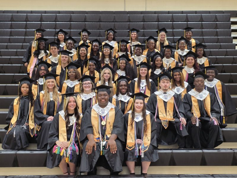 A large group of graduates in caps and gowns pose together on bleachers, smiling for a group photo in an indoor setting.