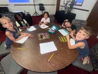 Five children sit around a table drawing with colored pencils and crayons on white paper in a classroom setting.
