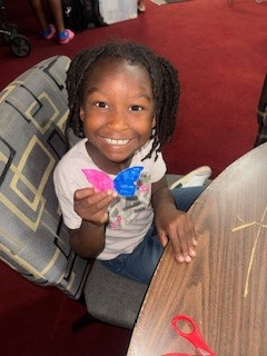 Smiling child holding a colorful paper butterfly while sitting at a table with craft materials on a red carpeted floor.