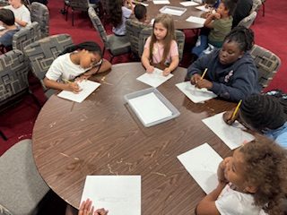 A group of children sit around a round table, drawing on white paper with pencils in a classroom or activity setting.