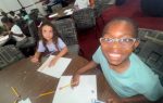 Smiling child with glasses holding a pencil at a table with paper, another child beside them, classroom setting with several children in the background.