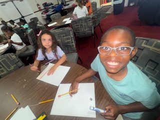 Smiling child with glasses holding a pencil at a table with paper, another child beside them, classroom setting with several children in the background.
