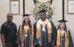 Four people pose in front of a banner reading "Dillon" with a wildcat logo; three are wearing graduation caps and gowns, and one is dressed in black polo attire.