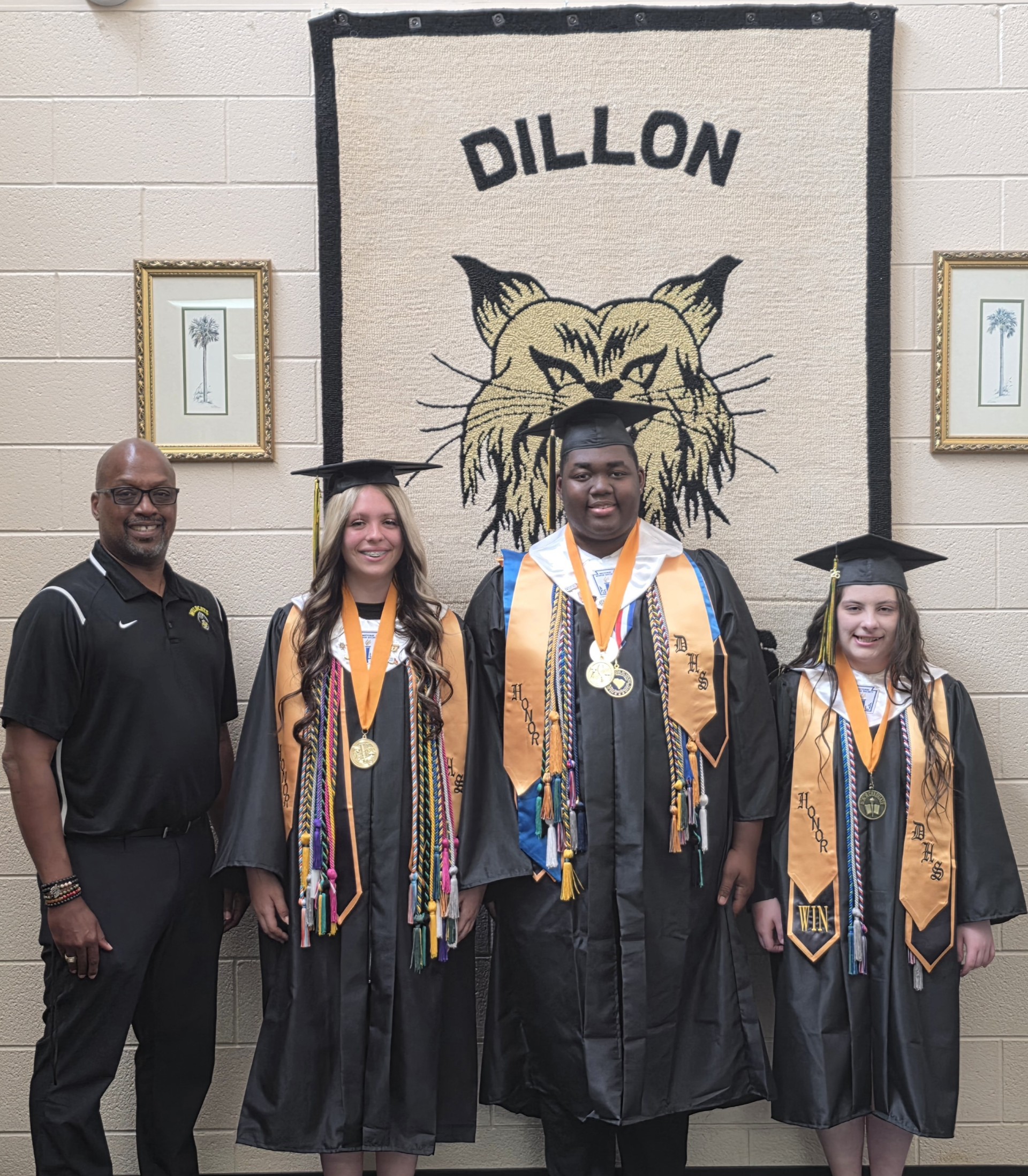 Four people pose in front of a banner reading "Dillon" with a wildcat logo; three are wearing graduation caps and gowns, and one is dressed in black polo attire.