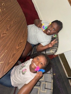 Two smiling children sitting at a table, holding colorful objects to their ears and flashing peace signs, looking up at the camera.