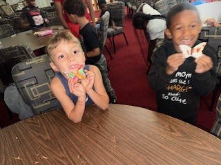 Two smiling young boys sit at a table, holding decorated cookies, in a room with other children and patterned chairs in the background.