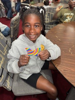 Smiling girl sitting at a table, holding a colorful, rainbow-striped craft shaped like a bird, in a busy indoor setting with other children in the background.